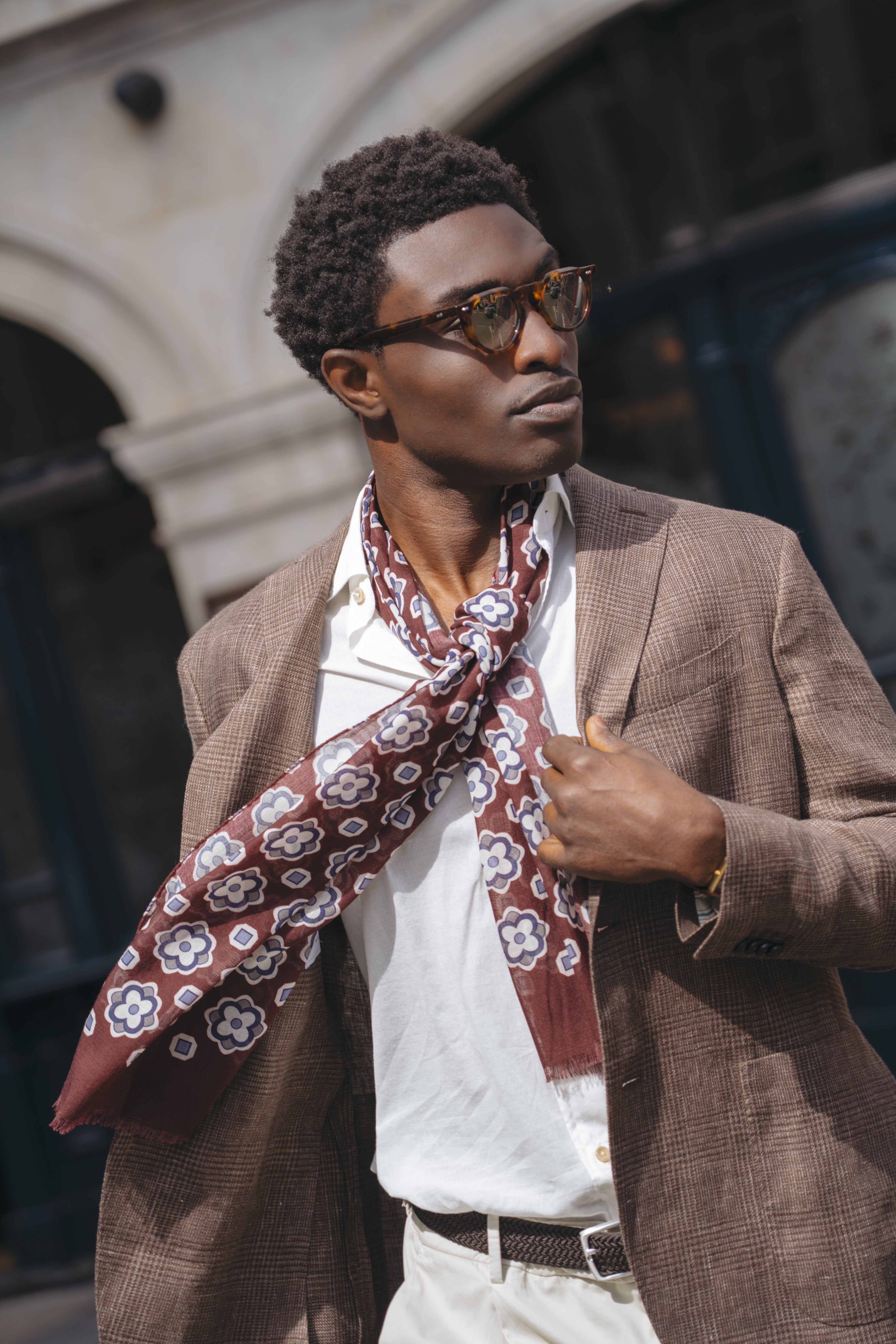 A confident man in sunglasses wears a Boglioli Brown Prince Of Wales Check Linen Jacket over a white shirt with a maroon floral scarf, standing outside by stone architecture and dark window panels, holding his blazer lapel. , available at Richard Gelding Mayfair