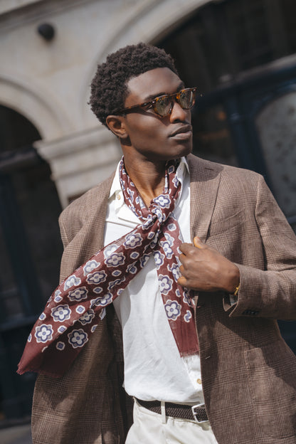 A confident man in sunglasses wears a Boglioli Brown Prince Of Wales Check Linen Jacket over a white shirt with a maroon floral scarf, standing outside by stone architecture and dark window panels, holding his blazer lapel. , available at Richard Gelding Mayfair
