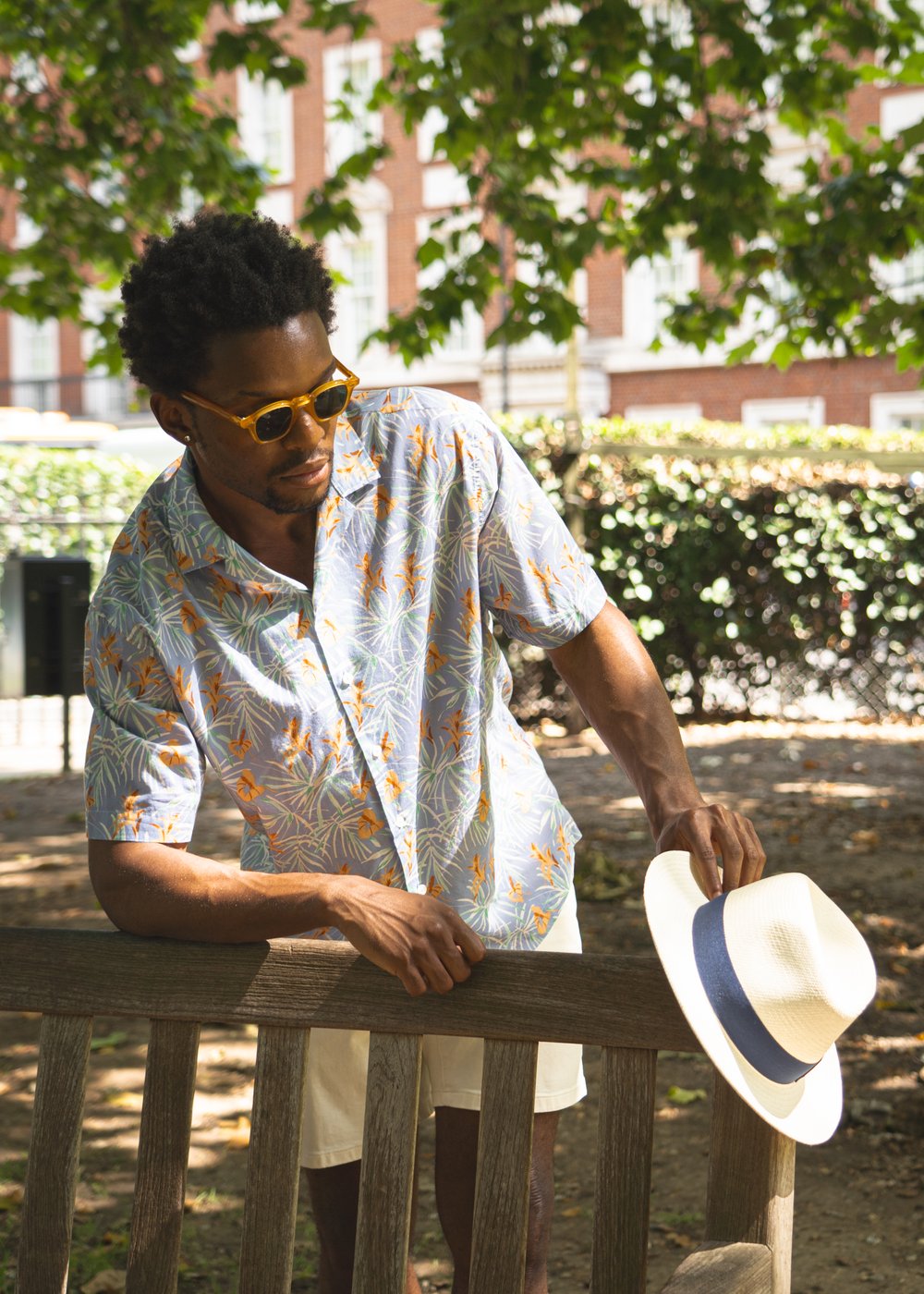 A man wearing sunglasses and a patterned short-sleeve shirt stands outdoors by a wooden bench, holding a white hat with a dark band. Sunlight filters through leafy trees, and a brick building is visible in the blurred background. , available at Richard Gelding Mayfair