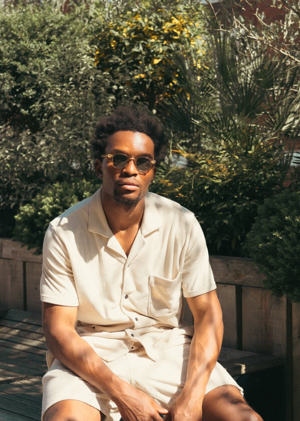 A man wearing sunglasses and a light beige short-sleeved shirt and shorts sits on a wooden bench outside, surrounded by lush green plants. Sunlight casts dappled shadows on him, and he gazes calmly toward the camera. , available at Richard Gelding Mayfair