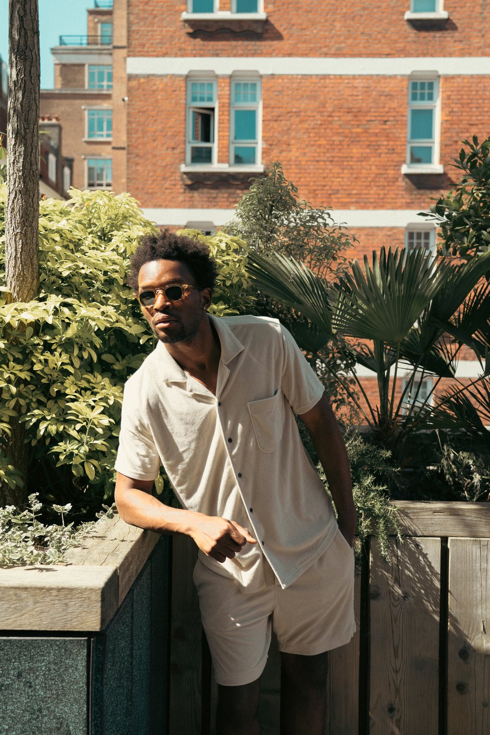 A man wearing sunglasses and a light beige short-sleeve shirt with matching shorts leans casually against a wooden railing. He is outdoors near lush green plants, with a brick building in the background under sunlight. , available at Richard Gelding Mayfair