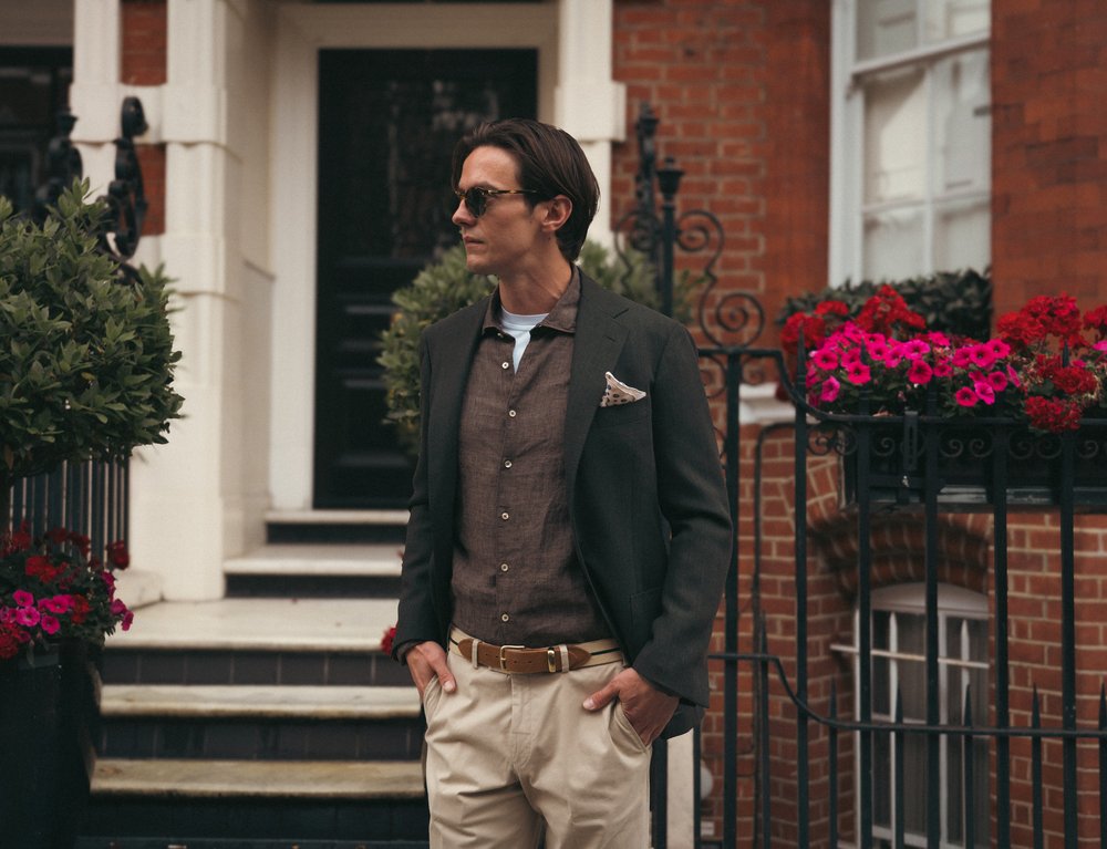 A man with short brown hair wears sunglasses, a dark blazer, brown shirt, beige pants, and a pocket square. He stands outdoors with hands in pockets in front of a brick building, surrounded by potted plants and vibrant pink flowers. , available at Richard Gelding Mayfair
