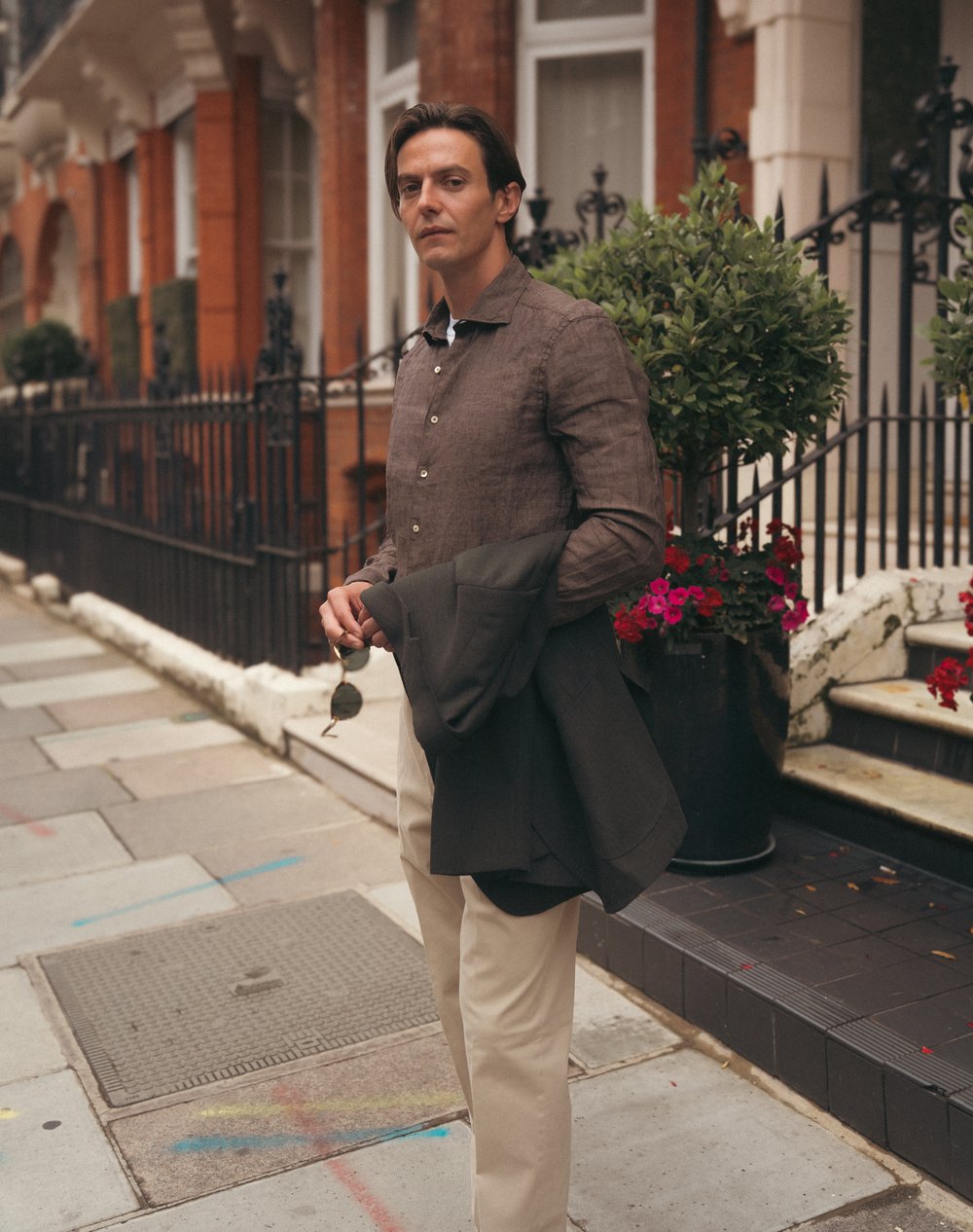 A man stands on a city sidewalk holding sunglasses and a dark jacket. He wears a brown shirt and beige pants. Behind him are red brick buildings, black railings, and potted plants with green leaves and red flowers. The scene looks calm and urban. , available at Richard Gelding Mayfair