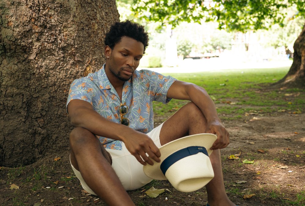 A man sits on the grass under a large tree, looking down thoughtfully at a white hat with a blue band in his hands. He wears a patterned short-sleeve shirt, white shorts, and sunglasses hanging from his shirt, with sunlight filtering through the leaves. , available at Richard Gelding Mayfair