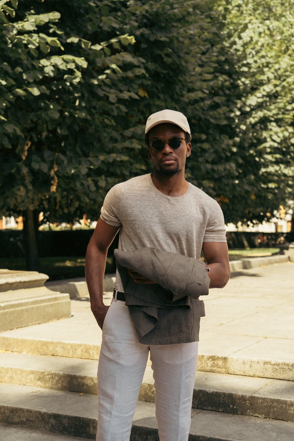 A man stands outdoors on stone steps, surrounded by leafy trees. He wears a beige cap, sunglasses, a light beige t-shirt, and white pants. He carries a folded taupe jacket in his arm, and sunlight filters through the trees, casting dappled shadows. , available at Richard Gelding Mayfair