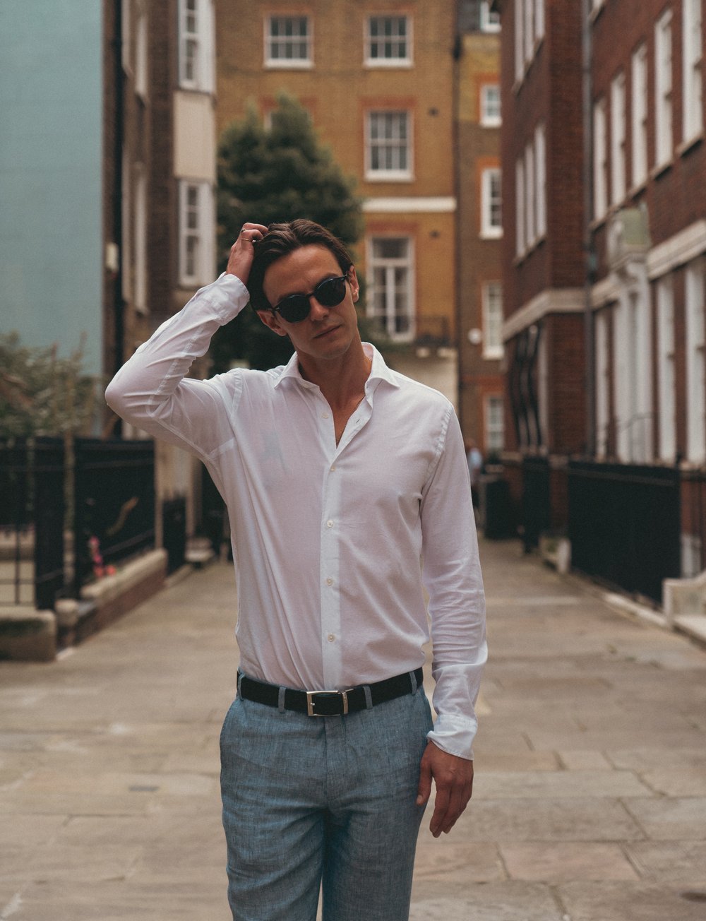 A man wearing sunglasses, a white button-down shirt, and light blue trousers stands on a narrow city street lined with brick buildings. He casually runs a hand through his hair, looking slightly to the side with a relaxed expression. , available at Richard Gelding Mayfair