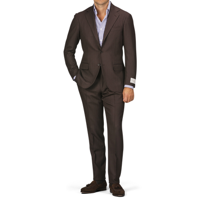 Person wearing a Brown Herringbone Calm Twist Wool Suit by Ring Jacket with a light blue dress shirt, standing with one hand in their pocket against a plain background.