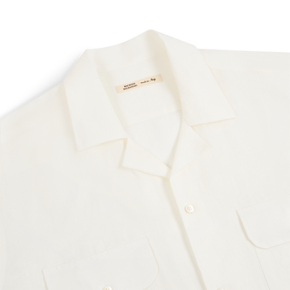 A close-up of the Maurizio Baldassari White Linen Army Shirt shows its short sleeves, notched collar, two buttoned chest pockets, and lightweight linen fabric. A beige inner tag displays the product name. The shirt is set against a plain white background. , available at Richard Gelding Mayfair