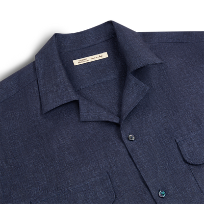 The Maurizio Baldassari Denim Blue Linen Army Shirt, featuring a textured fabric, two chest pockets, and black buttons, is neatly folded with its inner tag visible and laid flat on a white background. , available at Richard Gelding Mayfair