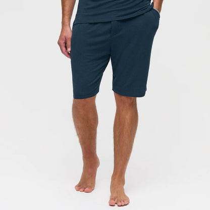 A barefoot man stands against a plain white background, wearing Homebody Navy Modal Jersey Pyjama Shorts and a matching shirt—ideal for luxe bedtime comfort. His left hand is in his pocket, and his right arm hangs by his side, face unseen. , available at Richard Gelding Mayfair