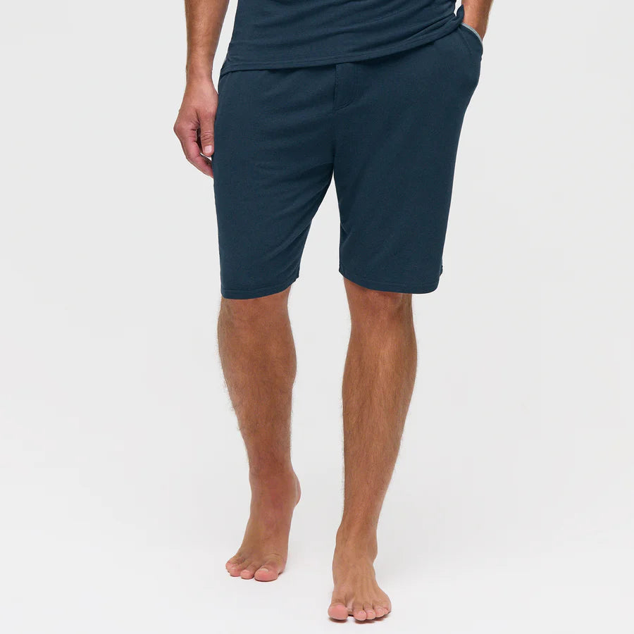 A barefoot man stands against a plain white background, wearing Homebody Navy Modal Jersey Pyjama Shorts and a matching shirt—ideal for luxe bedtime comfort. His left hand is in his pocket, and his right arm hangs by his side, face unseen. , available at Richard Gelding Mayfair
