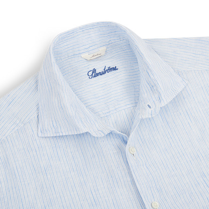A close-up of the Stenströms Blue Stripe Linen Slimline Shirt, neatly folded on a white background. The shirt features light blue and white stripes, a pointed collar with the brand label, and subtly textured linen fabric. , available at Richard Gelding Mayfair