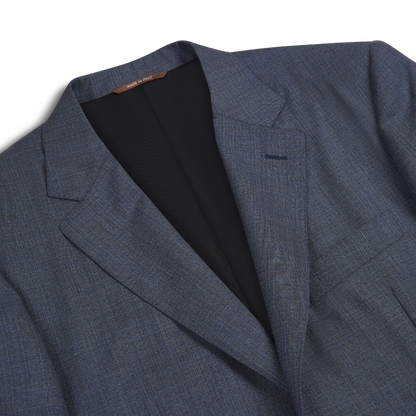 A close-up of the Canali Blue Texture Design Wool Suit shows its subtle plaid pattern, notched lapel, chest pocket, and black lining. The "Made in Italy" collar tag is visible. The textured suit is laid flat on a white background. , available at Richard Gelding Mayfair