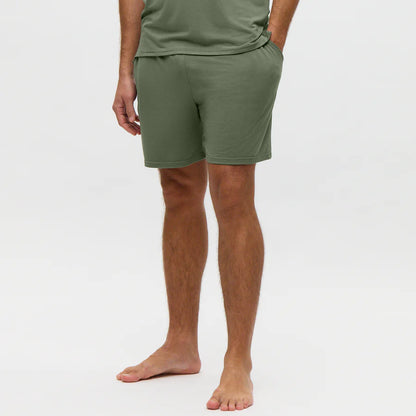 A person stands barefoot on a plain light background, hands in their pockets, wearing Homebody’s Charred Green Modal Wimbledon Shorts. The relaxed fit shorts are the focus, with the image cropped at shoulders and feet. , available at Richard Gelding Mayfair
