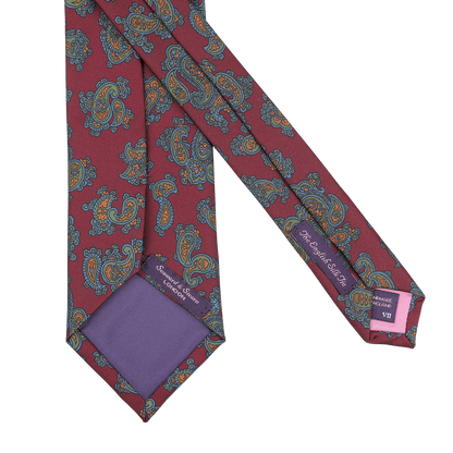 This Burgundy Paisley Printed Silk Tie by Seaward & Stearn is a handmade silk masterpiece. It features a red paisley design accentuated with blue and orange patterns, complemented by a luxurious purple lining and a discreet logo on the inner side.