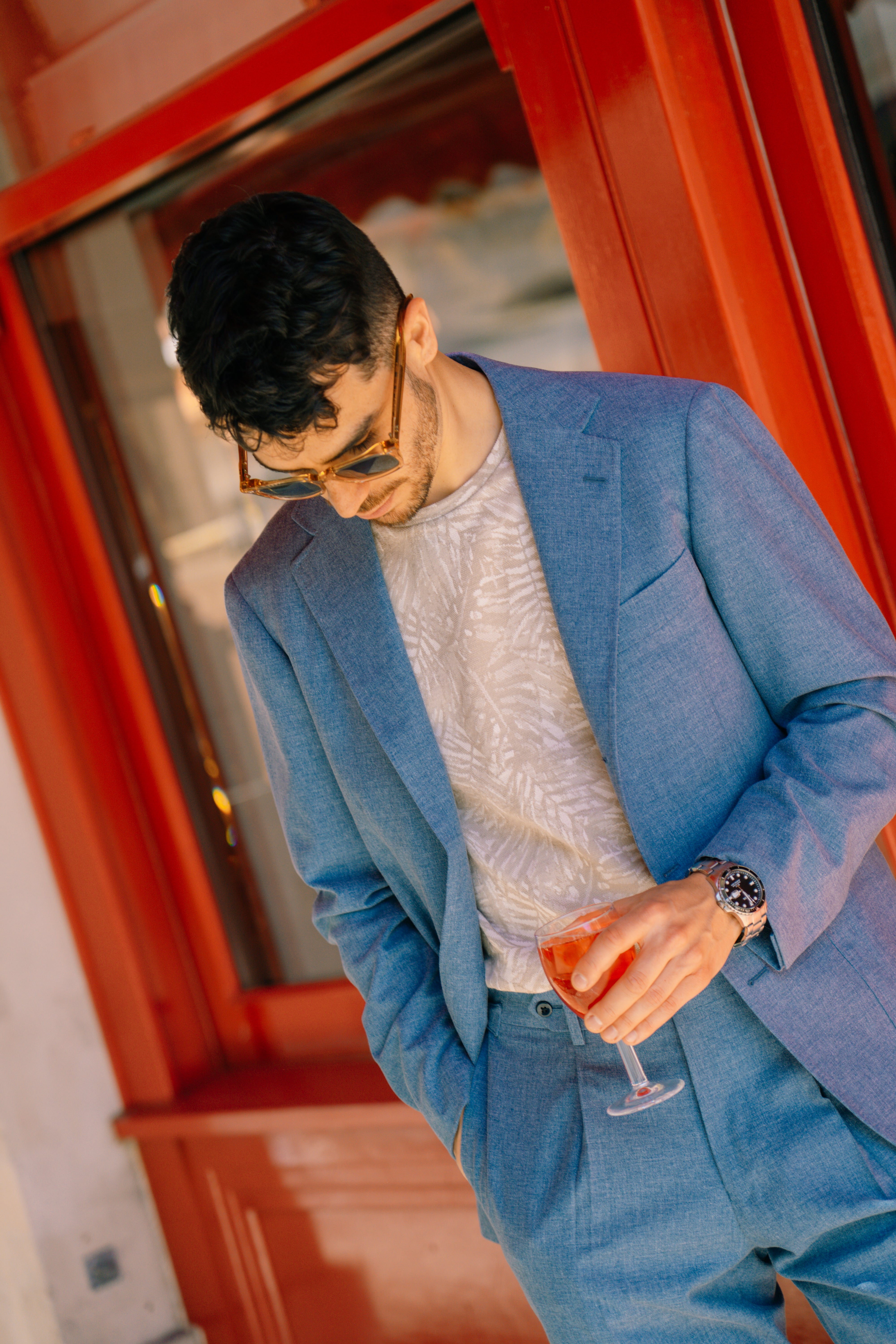 A man in a light blue suit and Maurizio Baldassari Beige Silk/Cotton Palm Tree Print T-Shirt stands by a red wall, wearing sunglasses and a watch, holding an orange drink in a wine glass as sunlight casts a warm glow over the scene. , available at Richard Gelding Mayfair
