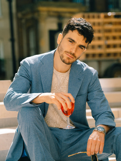 A man with short dark hair and a trimmed beard sits outdoors in sunlight, wearing the Ring Jacket Pale Blue Melange Tropical Wool/Cotton/Silk Suit over a patterned beige shirt. He holds sunglasses and a glass of red drink, looking calm. Blurred steps and a building are behind him. , available at Richard Gelding Mayfair