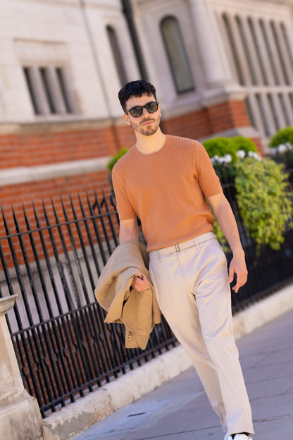 A man with short dark hair and sunglasses walks on a city sidewalk wearing a Gran Sasso Rust Chevron Weave T-Shirt, beige trousers with a belt, and white sneakers. He carries a tan jacket. A black iron fence and greenery are behind him. , available at Richard Gelding Mayfair