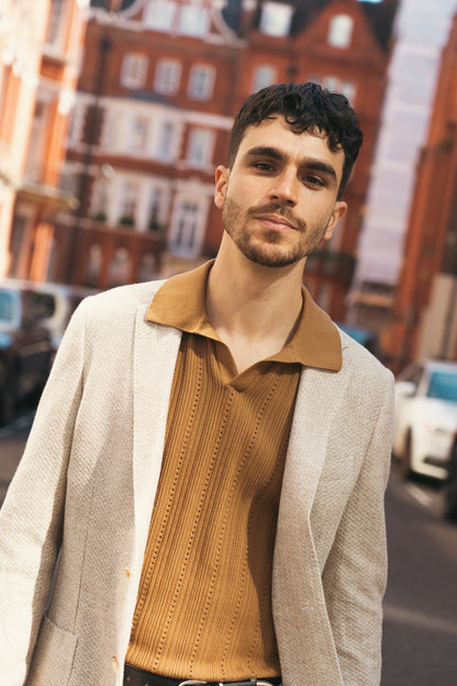 A man with short dark hair and a trimmed beard wears the Maurizio Baldassari Beige Linen Knitted Jersey Travel Blazer over a brown collared shirt, standing confidently on a sunlit street with red brick buildings in the background. , available at Richard Gelding Mayfair