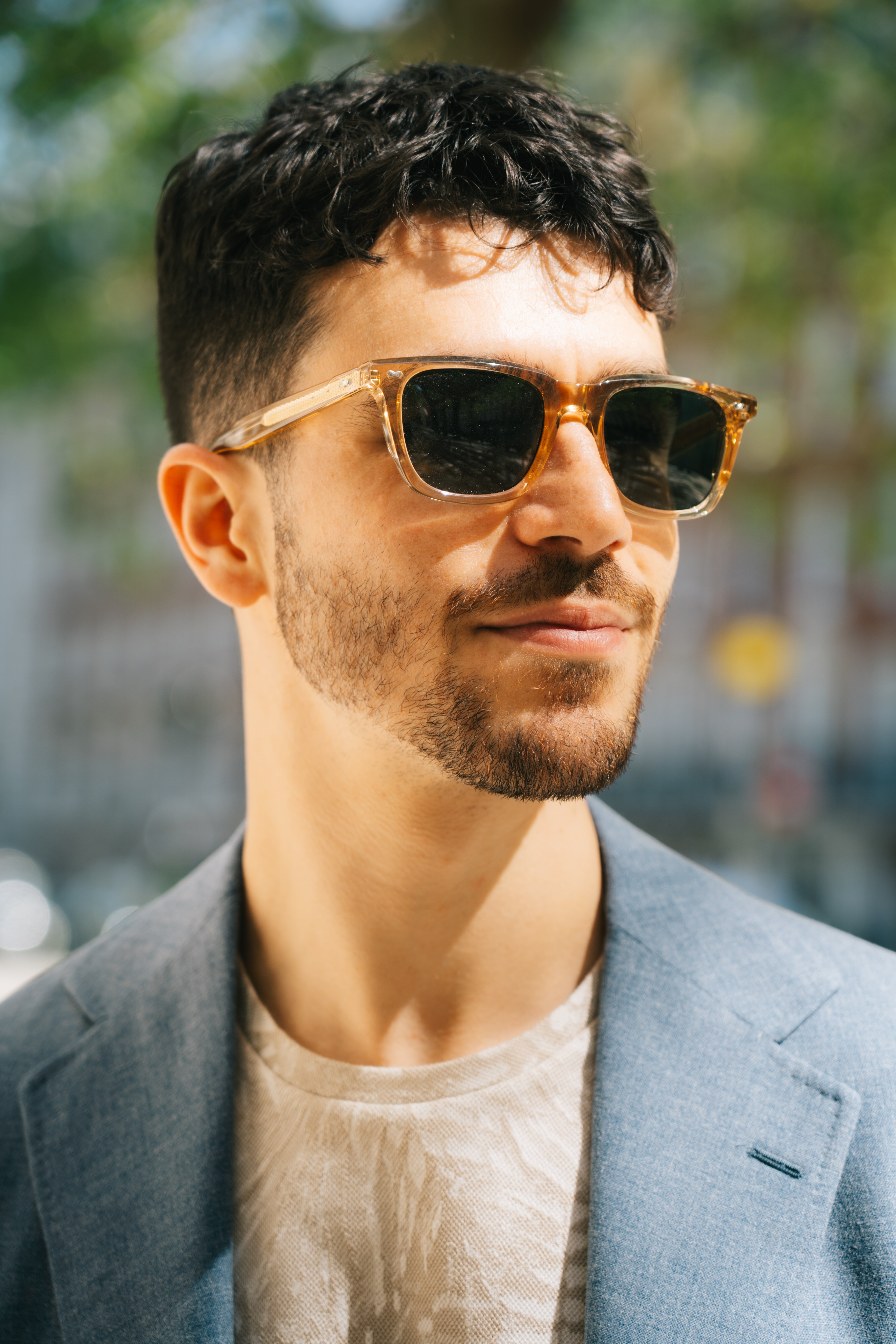 A young man with short dark hair and a trimmed beard wears TBD Eyewear’s Carré Eco Champagne | Bottle Green sunglasses and a light blue blazer over a cream shirt while standing outdoors in the sunlight. , available at Richard Gelding Mayfair