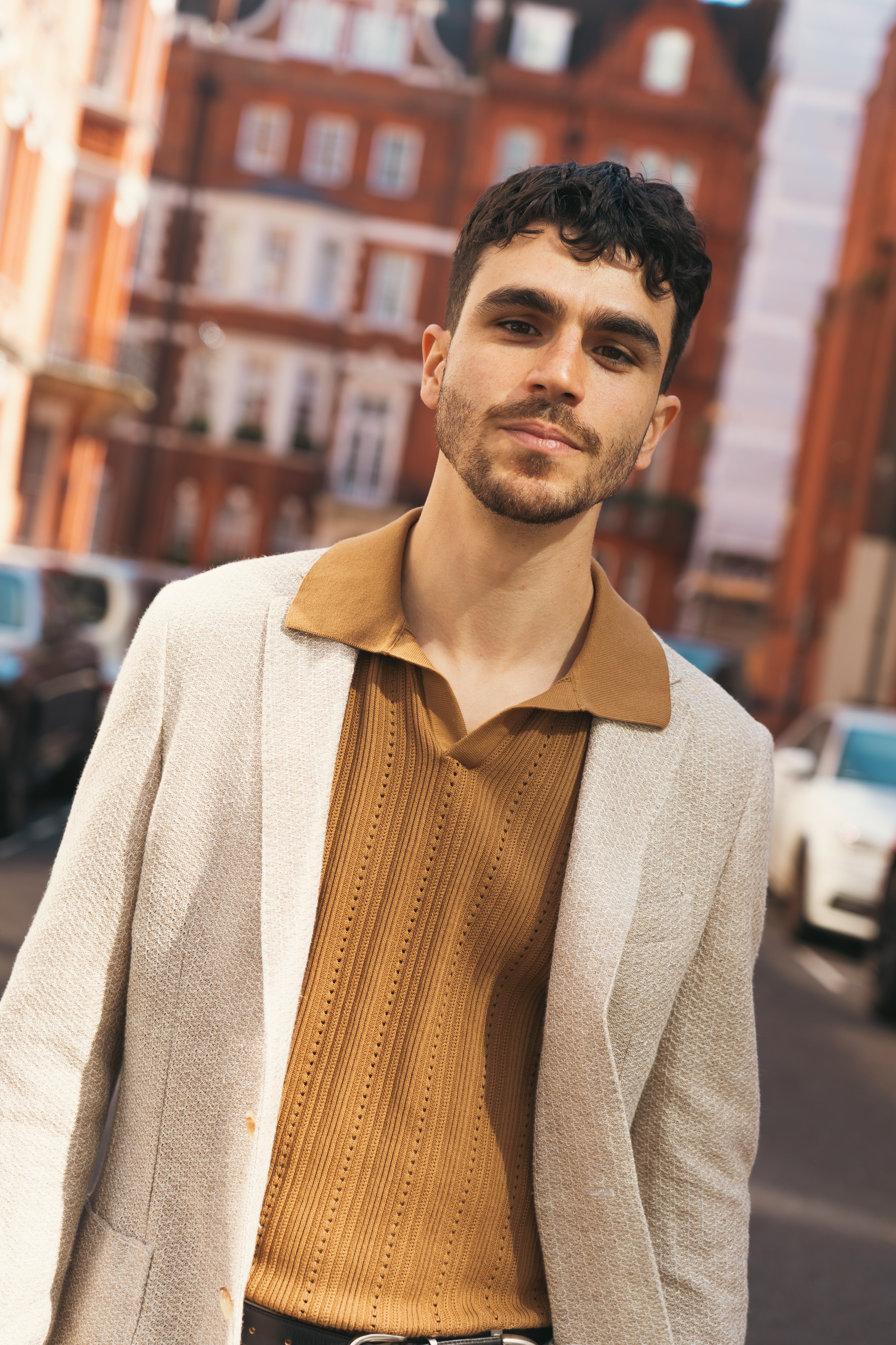 A confident young man stands on a city street, wearing Altea's Caramel Brown Diamond Cotton Polo. His dark hair and trimmed beard are complemented by blurred red-brick buildings and parked cars in the background. , available at Richard Gelding Mayfair