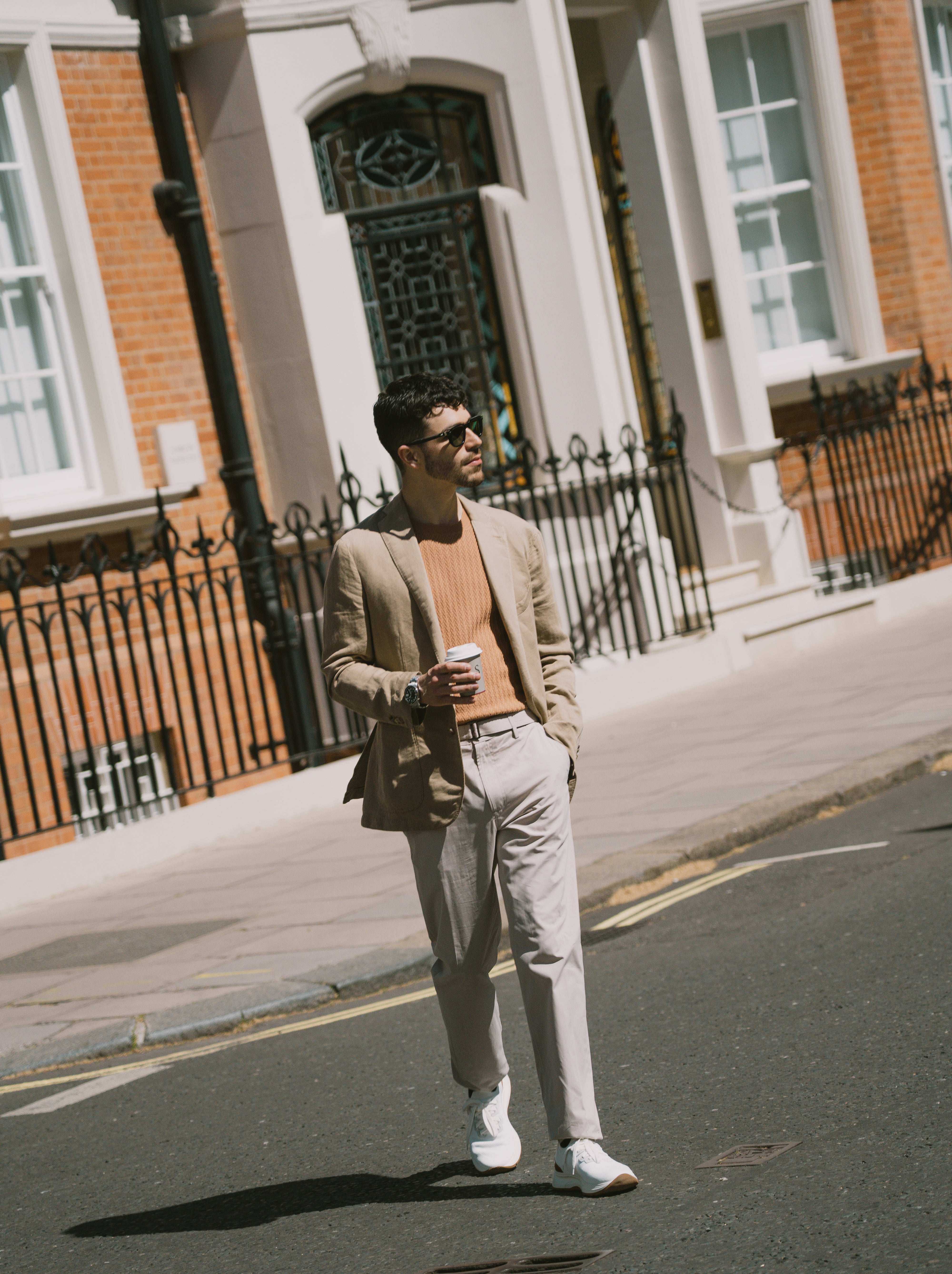 A man with dark hair and sunglasses walks down a city street holding a coffee, wearing the Boglioli Camel Beige Satin Cotton/Linen Jacket, a light orange sweater, light pants, and white sneakers. Red brick buildings with white trim line the background. , available at Richard Gelding Mayfair