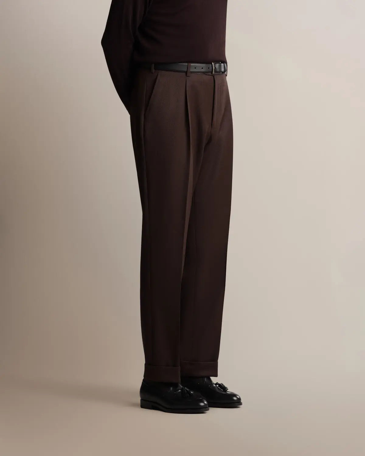 A person in profile wears Cavour Brown Straight Leg Pleated VBC Covert Wool Trousers with a thin black leather belt and black leather tassel loafers against a plain, light taupe background. , available at Richard Gelding Mayfair