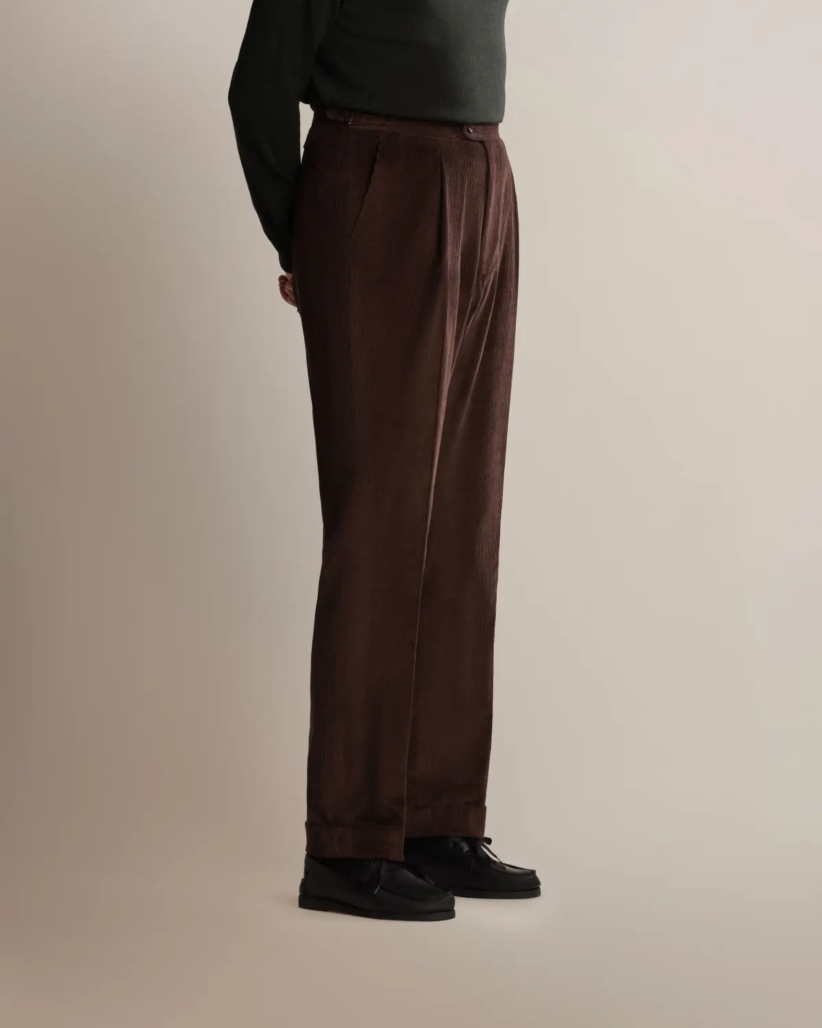 A person in profile wears Cavour Brown Wide Leg Pleated Corduroy Trousers with a dark green long-sleeved shirt and black shoes against a plain, light background. , available at Richard Gelding Mayfair