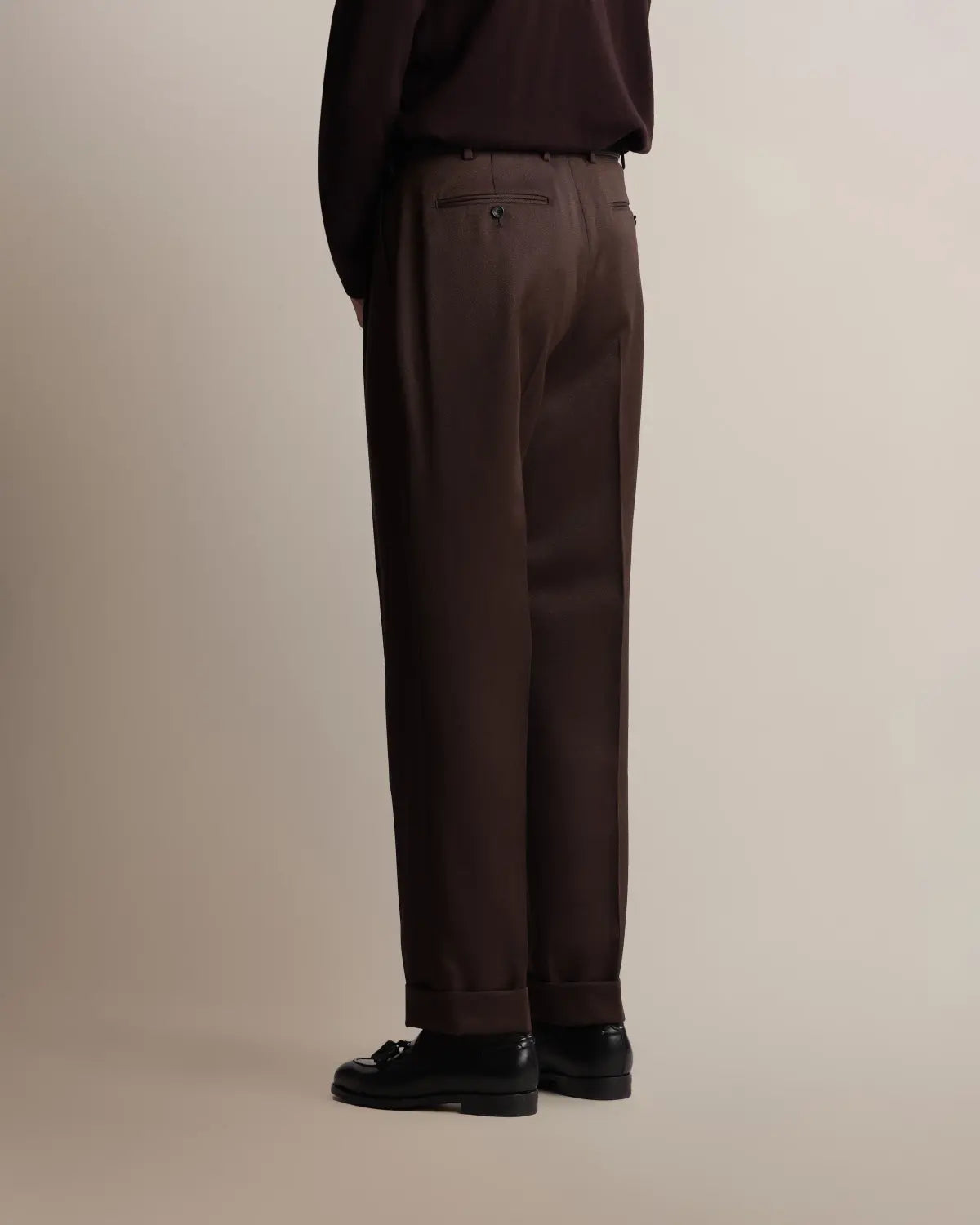 Seen from behind, a person wears brown straight-leg pleated VBC covert wool trousers by Cavour featuring a cuffed hem, double pleats at the waist, and two buttoned welt pockets, paired with a dark brown long-sleeve shirt and black leather loafers against a plain light background. , available at Richard Gelding Mayfair