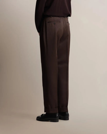 Seen from behind, a person wears brown straight-leg pleated VBC covert wool trousers by Cavour featuring a cuffed hem, double pleats at the waist, and two buttoned welt pockets, paired with a dark brown long-sleeve shirt and black leather loafers against a plain light background. , available at Richard Gelding Mayfair