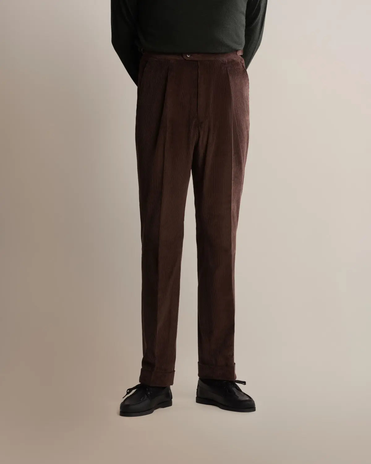 A person wears Cavour Brown Wide Leg Pleated Corduroy Trousers with deep front pleats and a cuffed hem, paired with a dark green long-sleeved shirt and black dress shoes against a plain, light grey studio background. , available at Richard Gelding Mayfair
