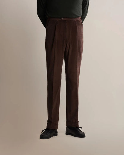 A person wears Cavour Brown Wide Leg Pleated Corduroy Trousers with deep front pleats and a cuffed hem, paired with a dark green long-sleeved shirt and black dress shoes against a plain, light grey studio background. , available at Richard Gelding Mayfair