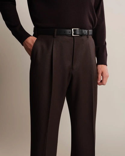 A person wears Cavour Brown Straight Leg Pleated VBC Covert Wool Trousers in dark brown, featuring a pleated front, held by a black leather belt with a silver buckle, and paired with a matching dark brown long-sleeved sweater, with one hand tucked into a side pocket against a plain background. , available at Richard Gelding Mayfair