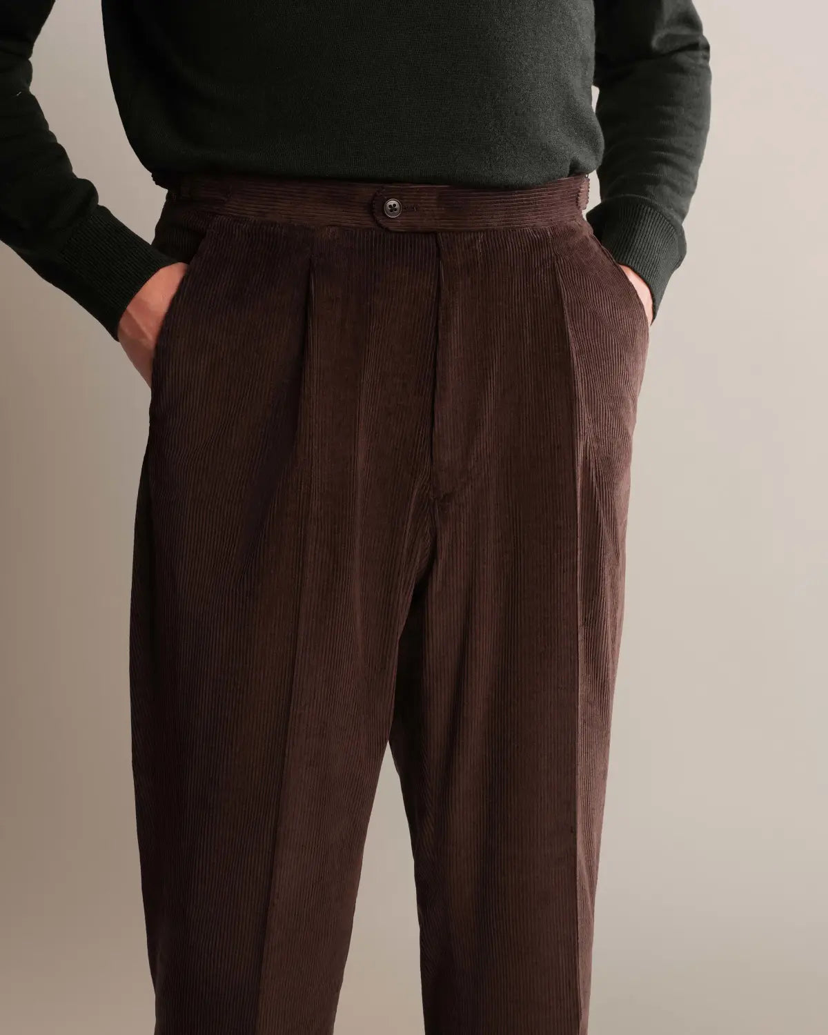 Cavour Brown Wide Leg Pleated Corduroy Trousers in dark brown, featuring a wide-leg, pleated fit and vertical ribbing, are worn with a dark green long-sleeved sweater; the person’s hands are tucked into the pockets against a light grey background. , available at Richard Gelding Mayfair