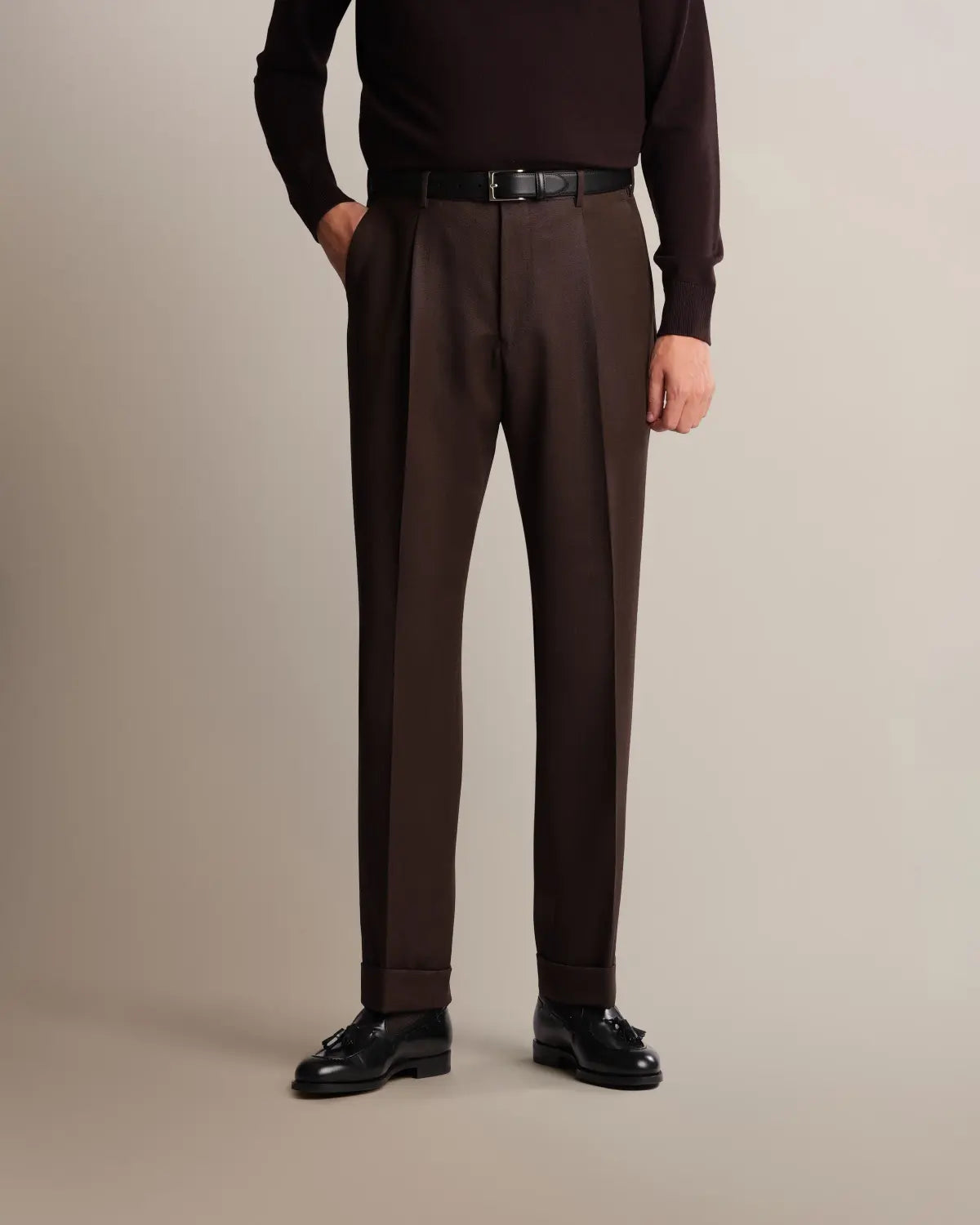A person wears Cavour Brown Straight Leg Pleated VBC Covert Wool Trousers in dark brown, featuring a straight-leg cut with a front pleat and cuffed hems, paired with a tucked-in dark brown sweater, a black leather belt, and black tassel loafers against a plain, light-toned background. , available at Richard Gelding Mayfair