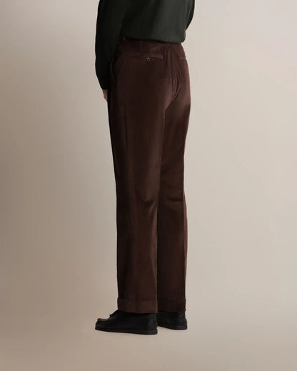 A person in profile wears Cavour Brown Wide Leg Pleated Corduroy Trousers tucked into a dark green long-sleeved shirt with black shoes against a plain light background. , available at Richard Gelding Mayfair