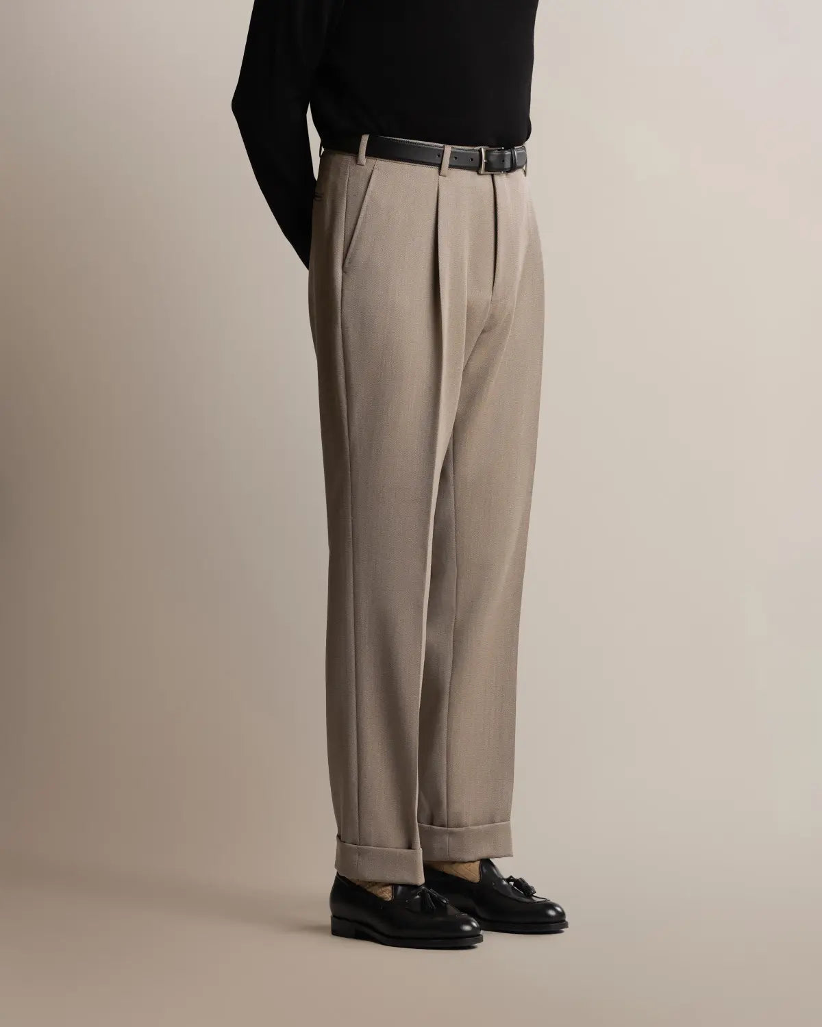 Cavour Taupe Straight Leg Pleated VBC Covert Wool Trousers with a pleated front and cuffed hems are worn with a black belt, black leather loafers, and a long-sleeved black top tucked in, viewed from the side against a neutral background. , available at Richard Gelding Mayfair