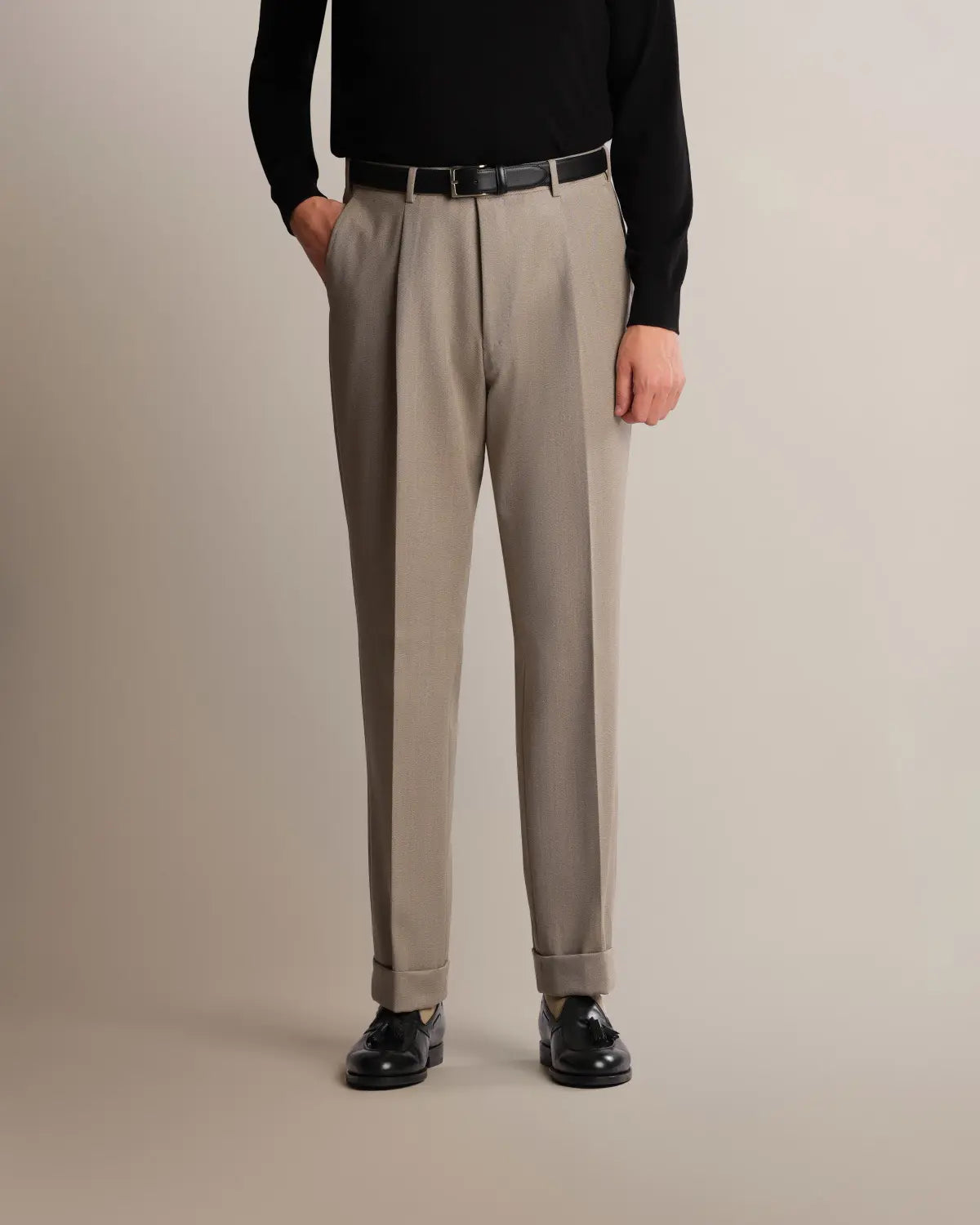 A man in a black sweater wears Cavour Taupe Straight Leg Pleated VBC Covert Wool Trousers with a sharp vertical crease, double pleats, and cuffed hems, paired with a black leather belt and tassel loafers against a solid, neutral light gray background. , available at Richard Gelding Mayfair