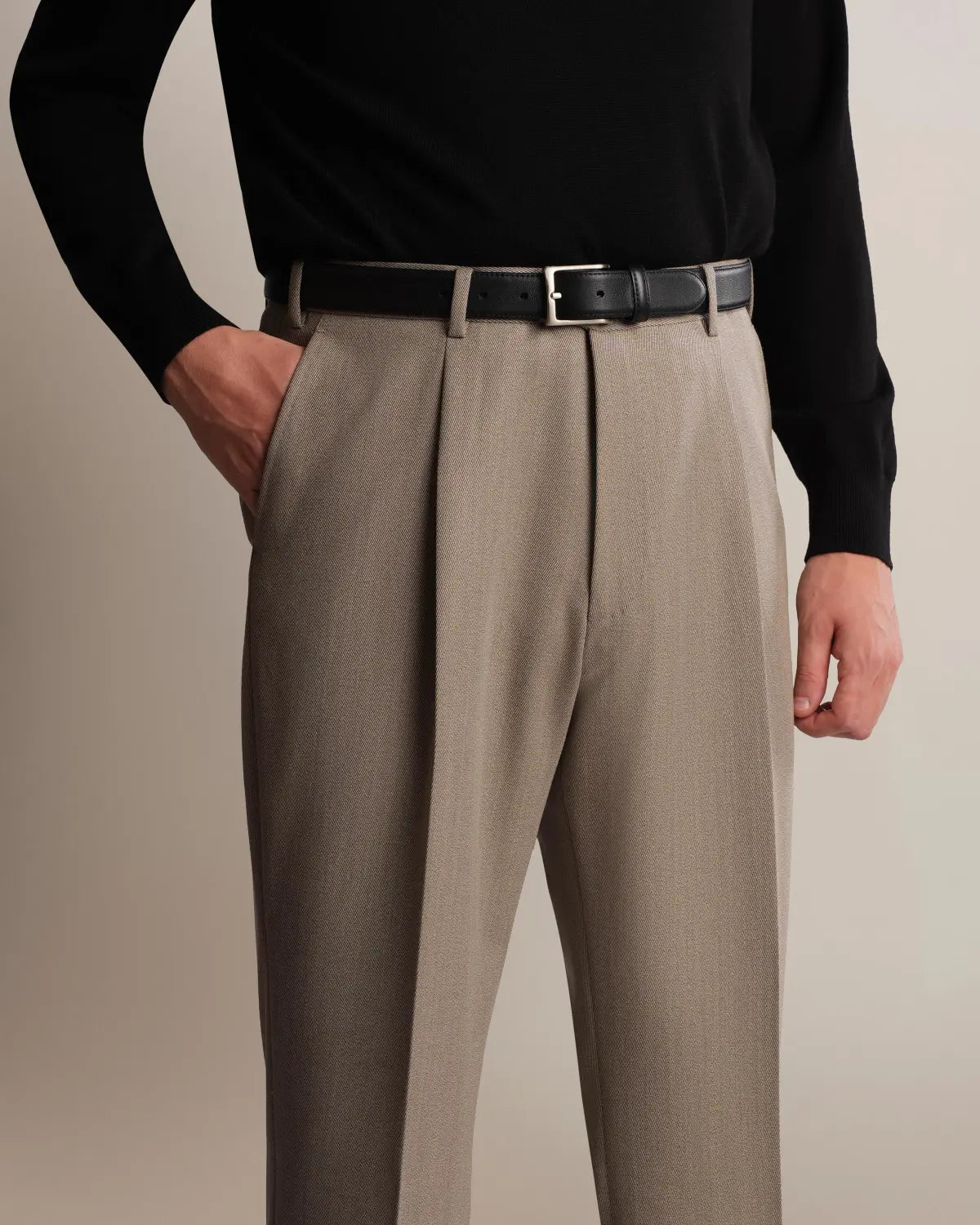 Cavour Taupe Straight Leg Pleated VBC Covert Wool Trousers in a fine, diagonal texture, worn with a black leather belt and long-sleeved sweater against a neutral background, with one hand tucked into the side pocket. , available at Richard Gelding Mayfair