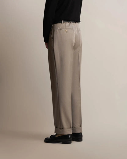 A person in profile wears Cavour Taupe Straight Leg Pleated VBC Covert Wool Trousers with a black long-sleeved shirt and black leather loafers against a plain, light gray background. , available at Richard Gelding Mayfair