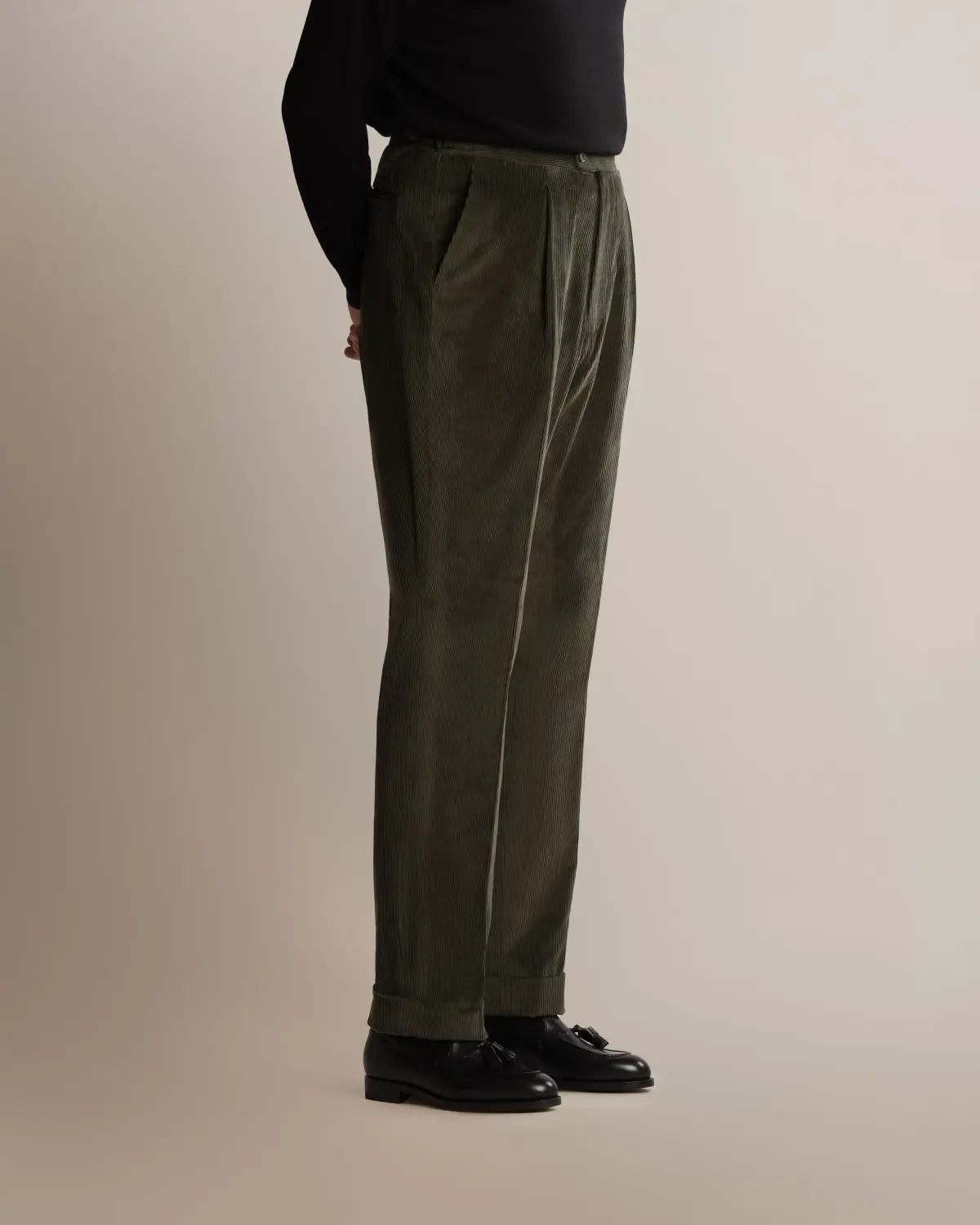 A side view of a person wearing Cavour Green Wide Leg Pleated Corduroy Trousers, featuring a high-waisted, wide-leg cut with a pleated waist and cuffed hem, paired with a black long-sleeved top and black tassel loafers. , available at Richard Gelding Mayfair