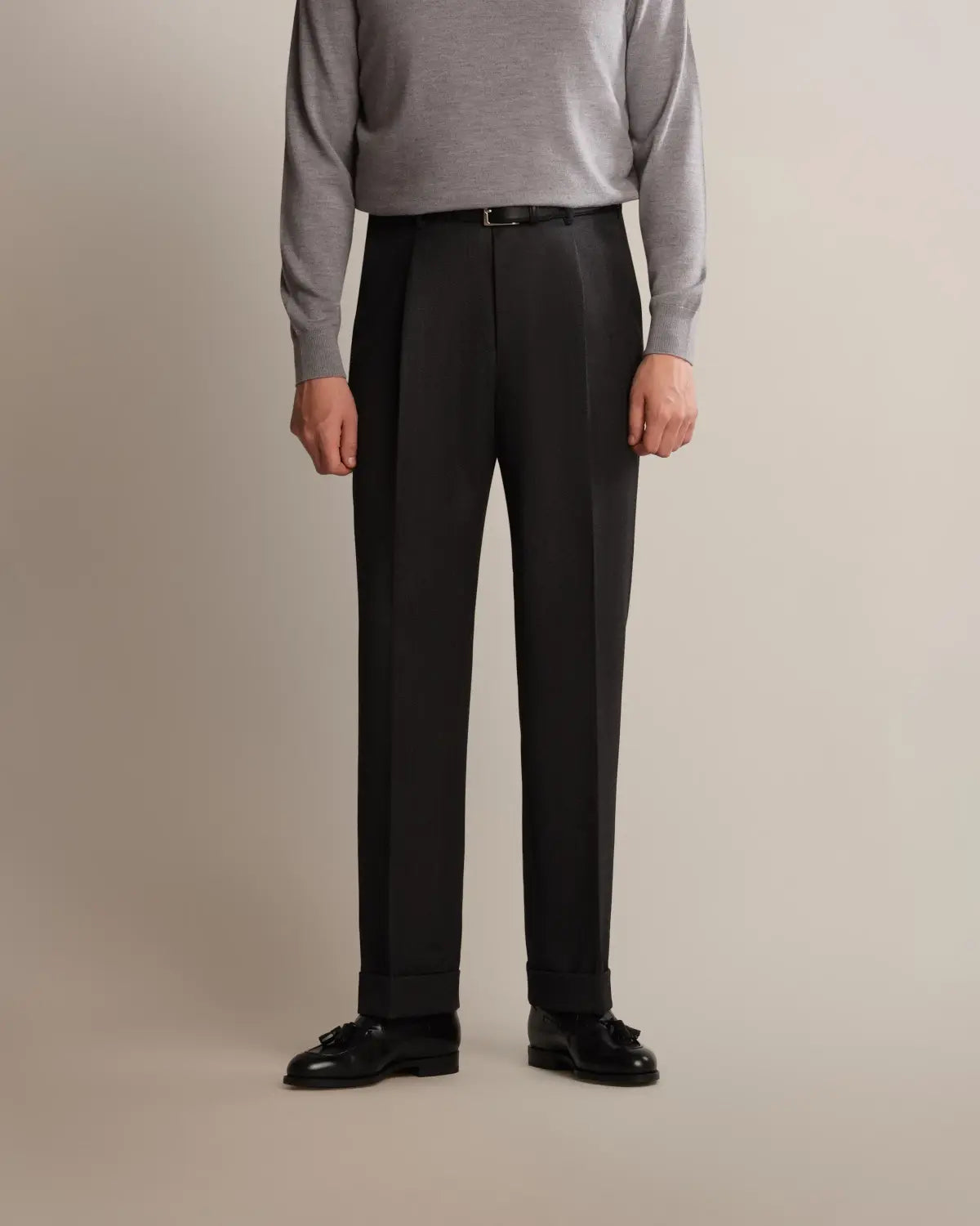 A person wears Cavour Charcoal Grey Straight Leg Pleated VBC Covert Wool Trousers with a visible front pleat and cuffed hems, paired with a tucked-in light grey crew-neck sweater, a thin black leather belt, and black leather tassel loafers against a neutral off-white background. , available at Richard Gelding Mayfair