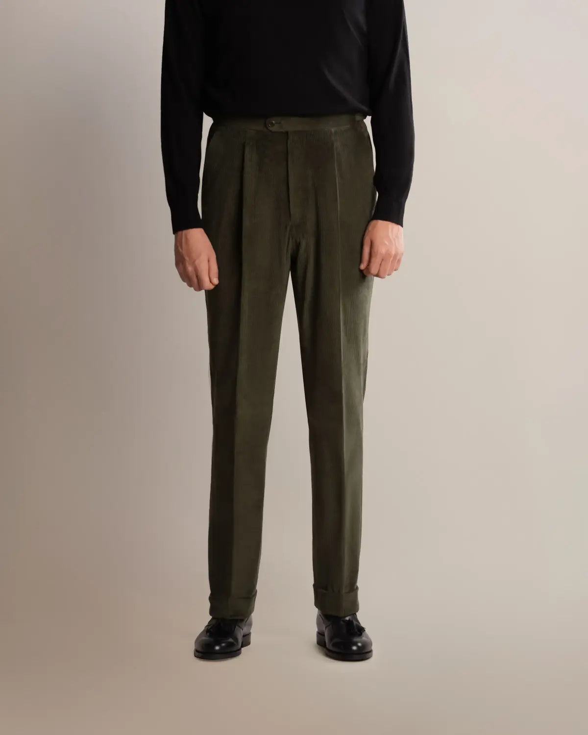 A person wears Cavour Green Wide Leg Pleated Corduroy Trousers with a wide-leg cut and cuffed hems, paired with a black long-sleeved sweater and black leather loafers against a plain, light grey background. , available at Richard Gelding Mayfair