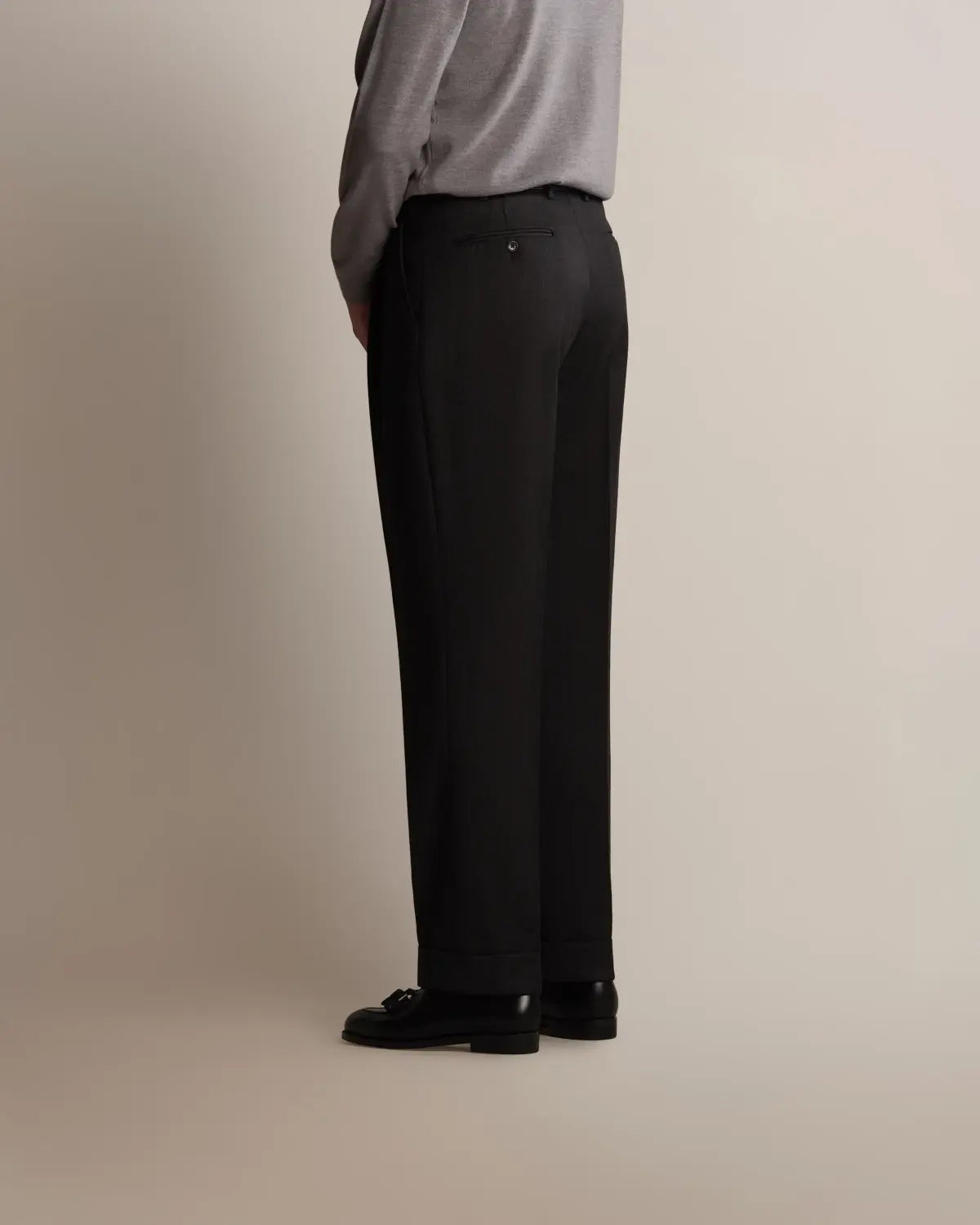 A side profile of a person wearing Charcoal Grey Straight Leg Pleated VBC Covert Wool Trousers with a cuffed hem, featuring a pleated front and buttoned back pocket, paired with a light grey long-sleeve top and black leather loafers against a plain light background. , available at Richard Gelding Mayfair