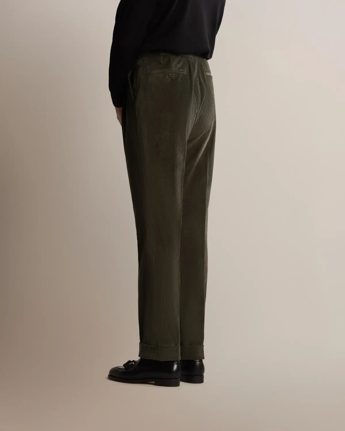 A person in profile wears Cavour Green Wide Leg Pleated Corduroy Trousers with a black long-sleeved top and black leather loafers against a plain, light beige background. , available at Richard Gelding Mayfair