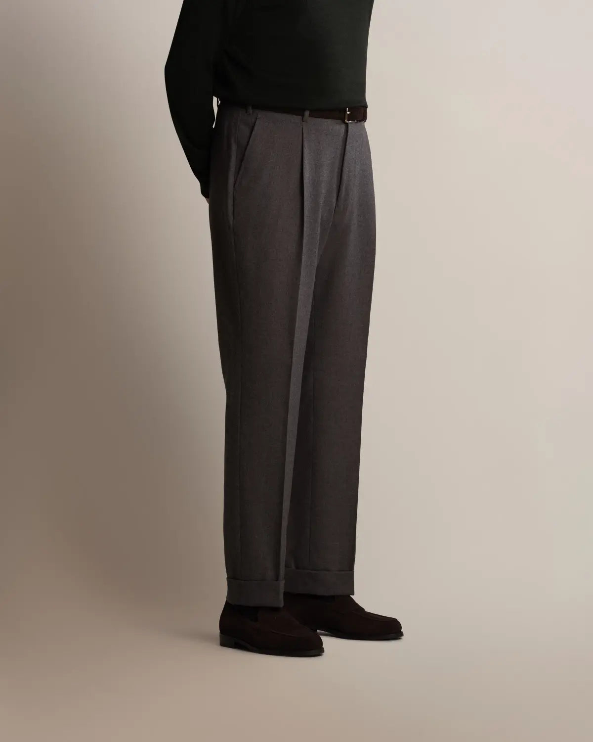 A person stands in profile wearing Cavour Grey Straight Leg Pleated Flannel Trousers, featuring charcoal grey fabric, a pleated front, and cuffed hems, paired with a dark brown belt, a black long-sleeved shirt, and dark brown suede loafers against a light background. , available at Richard Gelding Mayfair