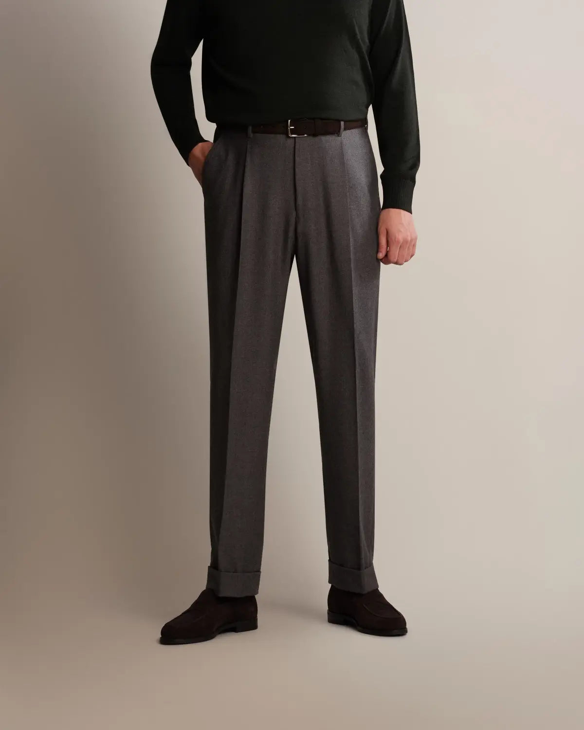A person wearing Cavour Grey Straight Leg Pleated Flannel Trousers with a slim brown belt, paired with a dark green long-sleeved sweater and dark brown suede shoes against a plain, light-toned background. , available at Richard Gelding Mayfair