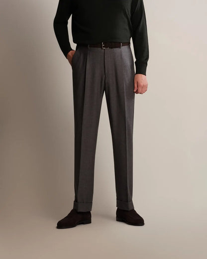 A person wearing Cavour Grey Straight Leg Pleated Flannel Trousers with a slim brown belt, paired with a dark green long-sleeved sweater and dark brown suede shoes against a plain, light-toned background. , available at Richard Gelding Mayfair