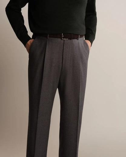 A person wears charcoal grey Cavour Grey Straight Leg Pleated Flannel Trousers, paired with a black long-sleeved sweater and a dark brown leather belt, with both hands tucked into the trouser pockets. , available at Richard Gelding Mayfair