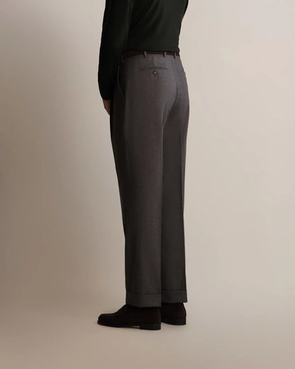 A person from the back wearing Cavour Grey Straight Leg Pleated Flannel Trousers with cuffed hems and a dark brown belt, styled with a tucked-in black long-sleeved shirt and dark brown suede shoes against a plain, light-toned background. , available at Richard Gelding Mayfair